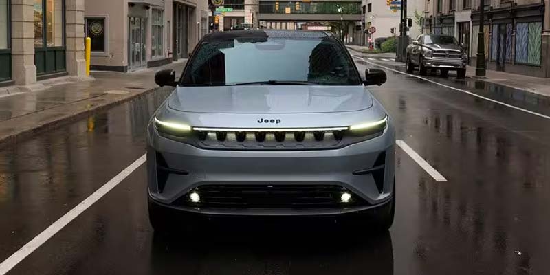 A silver Jeep Wagoneer parked on a wet city street with buildings in the background.